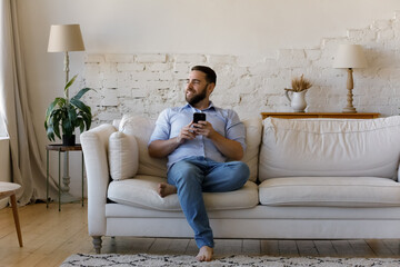 Smiling man sit on sofa at home with smartphone distracted from device look into distance spend...