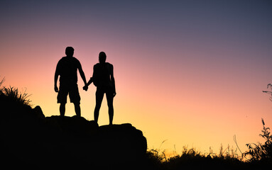 Couple hikers man and woman holding hands on top of a mountain looking out to the sunset view....