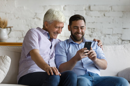 Older 50s man his grown up millennial son sit on sofa using smartphone spend weekend together at home. Diverse gen and modern tech, new mobile application usage, help with wireless device concept
