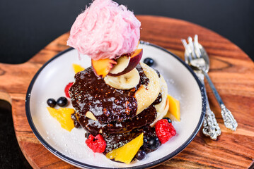 A serving of stacked pancakes with chocolate sauce, fruit pieces and pink fairy floss