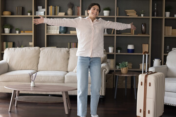 Full length overjoyed pretty young Hispanic woman having fun in modern living room, feeling excited coming back from interesting travel or waiting for entertaining vacation trip, dancing near suitcase