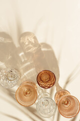 Set colored glasses from transparent and beige glass with beautiful shadows from palm leaf at sunlight, summer drink for events, white wine, water in trend glasses, aesthetic top view