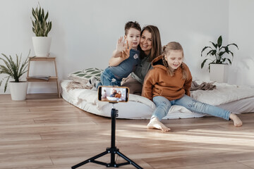 Obraz premium the family smiles and records a video on the phone. bloggers shoots a video clip at home. the phone is on a tripod. family mom and two children are sitting on the bed and posing for the camera