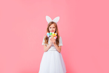 Obraz na płótnie Canvas Cute blonde girl 8-9 years old in a white T-shirt and skirt with rabbit ears holds Easter eggs isolated on a pink background. Happy Easter, holidays, traditions.