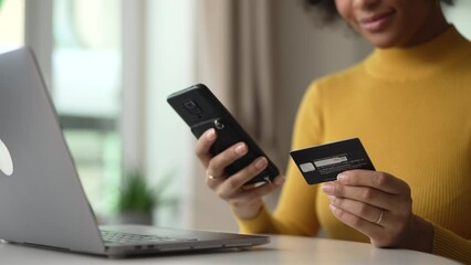 Young woman using wireless devices and credit card for online shopping in home room spbd. 4k video Close view of beautiful african female holds smartphone in hand and looks with smile, enters payment - Powered by Adobe