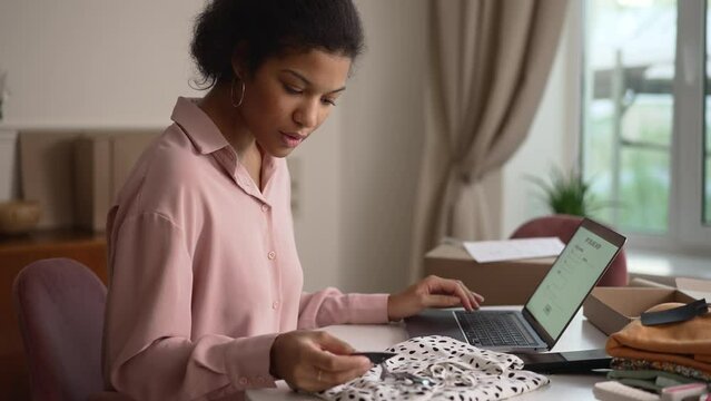 Young female client making return of clothes and sitting in front of laptop at table at home spbd. 4k video Close view of beautiful African female checks clothing and looks at computer screen, fills