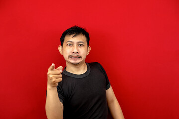 Face scolding Asian man in black t-shirt pointing at his hand. red background.