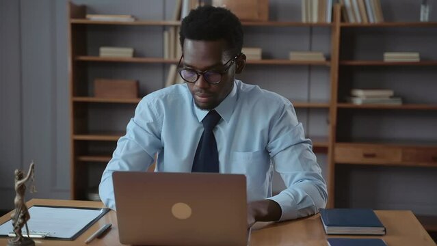 Male professional lawyer working with laptop and sitting at table in modern office spbas. Portrait of young American African man watches web on computer screen or does work and sits at desk in light