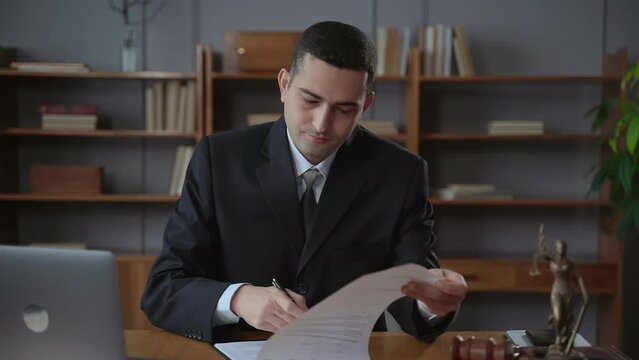 Successful businessman signing contract and sitting at table with laptop in modern office spbas. Portrait of young caucasian man reads conditions and signs paper document, looks with smile and sits at