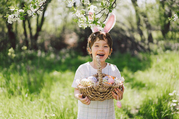 Easter egg hunt in spring garden. Funny boy with eggs basket and bunny ears on Easter egg hunt in...