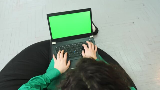 A girl sits on a cushioned chair and works with a green screen laptop. The green screen on the laptop. High quality 4k footage