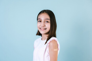 Hispanic girl child portrait after getting a vaccine protection and showing her arm with bandage receiving vaccination on blue background in Mexico Latin America
