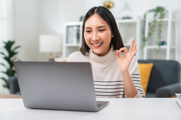 Cheerful beautiful Asian woman using laptop computer with showing ok sign and talk to camera at video calling recording with online chat at house.