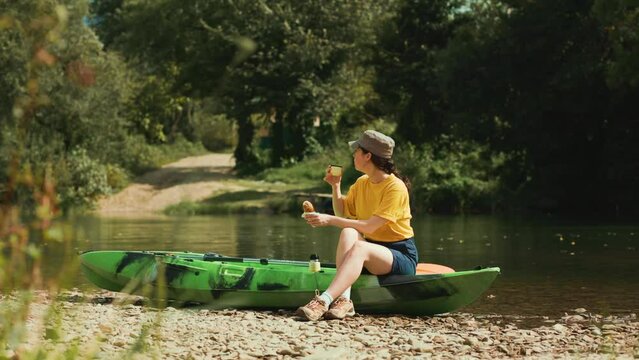 Sport and recreation. A young woman is sitting on a kayak and eating a hot dog, washing down a drink from a thermos. The concept of outdoor activities.