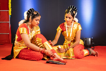 Friend helping while Bharatnatyam dancer suffering from leg or ankle injury while dancing on stage - concept of traditional dancers, togetherness and safety.