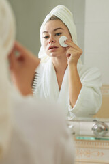 Young woman in bathrobe wiping face with cotton pad soaked in toner to restore skins pH level