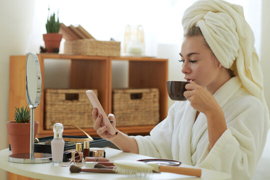 Young woman in bathrobe sitting at vanity, drinking coffee and reading news on smartphone