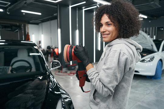 Pleased auto detailer with car polisher standing beside automobile