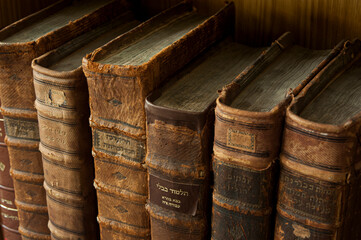 Dusty, worn and faded leather-bound Jewish holy books stacked upright on a library shelf. - obrazy, fototapety, plakaty