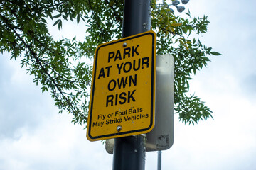 Low angle view of a Park At Your Own Risk sign outside of a baseball field, warning anyone parking their cars of the chance of fly balls