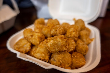 Selective focus of crispy, golden brown tater tots in a white styrofoam to-go container from a restaurant.