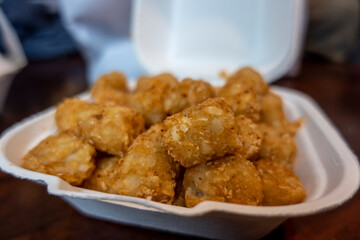 Selective focus of crispy, golden brown tater tots in a white styrofoam to-go container from a restaurant.