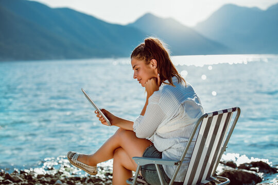 Smiling woman sitting on deck chair by the sea using tablet on a sunny day