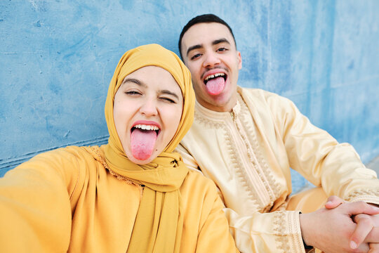 Young woman and man sticking out tongue in front of blue wall