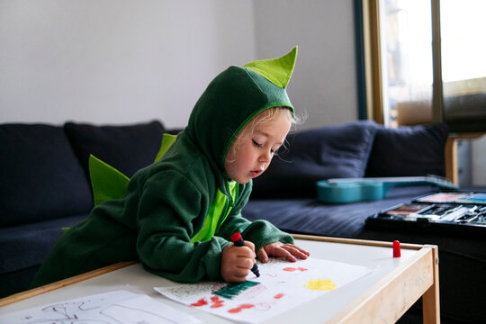 Boy in dinosaur costume drawing on paper in living room