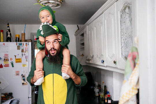 Father carrying boy on shoulders with dinosaur costume in kitchen at home