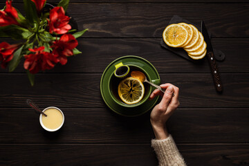 Woman in a cozy beige sweater drinking a big cup of hot tea with lemon and honey, on wooden background, overhead shot