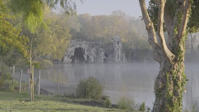 Artificial grotto at Majolan Park lake in Blanquefort France behind trees on misty morning, Dolly left shot