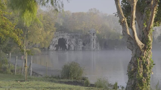 Concrete grotto cave at Majolan Park lake in Blanquefort France behind trees on misty morning, Locked shot