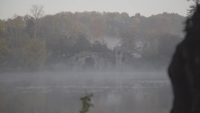 Grotto cave behind misty clouds lifting from Majolan Park lake in Blanquefort France, Locked shot