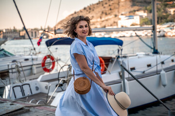 A young happy woman in a blue dress and hat stands near the seaport with luxury yachts. Travel and vacation concept