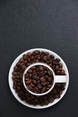 Fragrant coffee beans in a white coffee cup top view. Coffee beans on black slate copy space. Coffee beans in a cup on a black background flat lay.