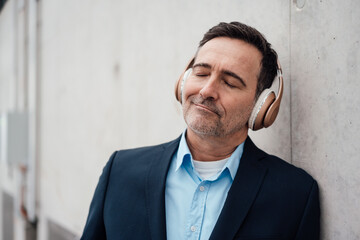Smiling businessman with eyes closed listening music through wireless headphones by wall