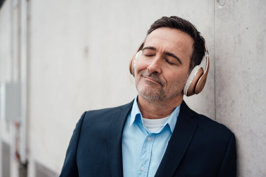 Smiling businessman with eyes closed listening music through wireless headphones by wall