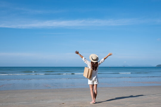 Rear view image of a woman with hat and bag raising hands while walking on the beach with blue sky background