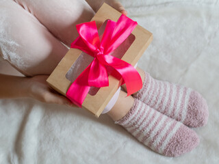 The hands of a young woman hold a box with a gift tied with a pink ribbon. A girl in light clothes sits on a white surface and holds a box with a gift.