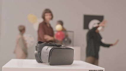 Slowmo close-up of virtual reality headset on stand in museum of contemporary arts with people examining art objects at exhibition in blurred background
