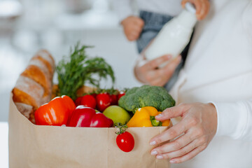 Young mother in white hoodie holding a baby girl and posing with paper package of fresh vegetables,...