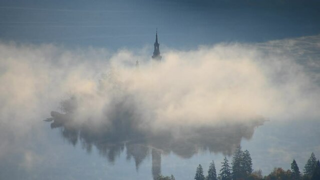Elevated time lapse of fog moving around small island on lake Bled, Slovenia. Aerial, telephoto view of famous Assumption of Maria Church tower. Fog or mist on the surface. Static shot