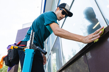 Fototapeta na wymiar A young male worker of a professional cleaning service in overalls washes the glass of the windows of the facade of the building. Clean showcases on request