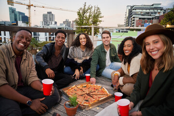 Group of diverse friends smiling at camera with red cups while sitting on a rooftop terrace in the city