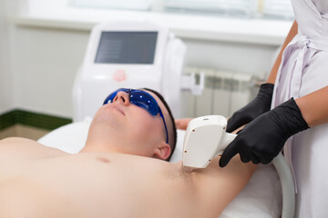 A young handsome man is lying on the couch of a cosmetology office in protective glasses with his hands raised during a laser hair removal procedure in the armpit
