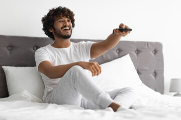 Joyful hindu guy sitting on bed, watching TV