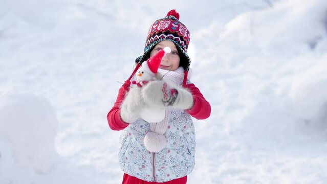 A little girl in warm, bright clothes holds a small snowman in her hands on a clear sunny winter day. Winter fun