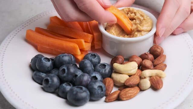 Woman hand eating healthy snack - carrots with peanut butter, berries and nuts on white plate. Healthy vegan food concept.