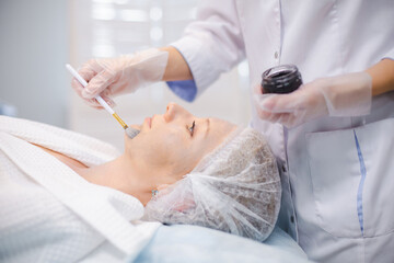 Obraz na płótnie Canvas Side view of hands of cosmetology specialist applying carbon nanogel on young woman patient face using brush, getting ready for carbon peeling procedures